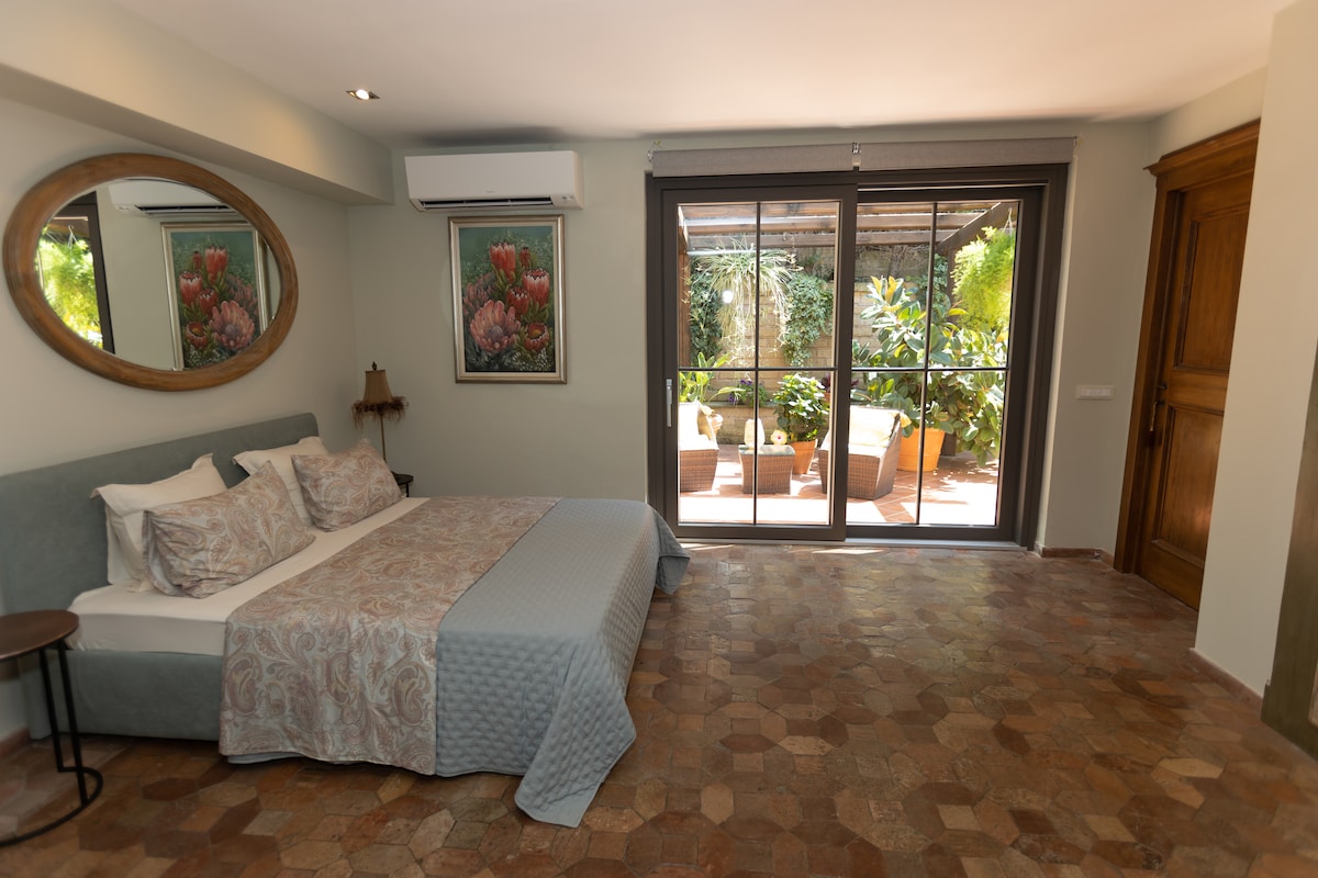A spacious room features a large bed with quality bedding and a stylish bedspread. A round mirror is mounted above a small side table. Glass doors lead to a terrace adorned with greenery and outdoor seating, allowing natural light to fill the space.