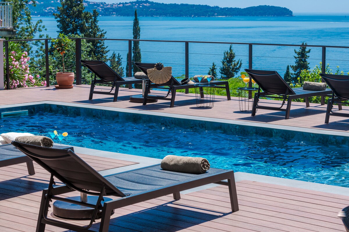 A private swimming pool glistens in the sunlight, surrounded by sun loungers equipped with rolled towels. Lush greenery and a sea view create a scenic backdrop, while the expansive deck provides ample space for relaxation.