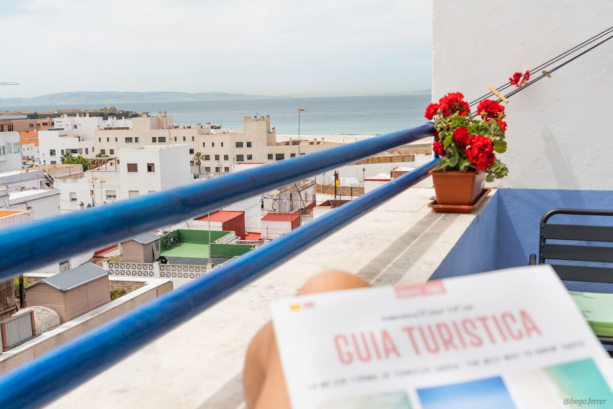 A terrace overlooks the city and sea, showcasing vibrant flowers in a pot along the railing. A travel guide is placed on the table, while colorful buildings and the ocean can be seen in the distance, contributing to a serene setting.