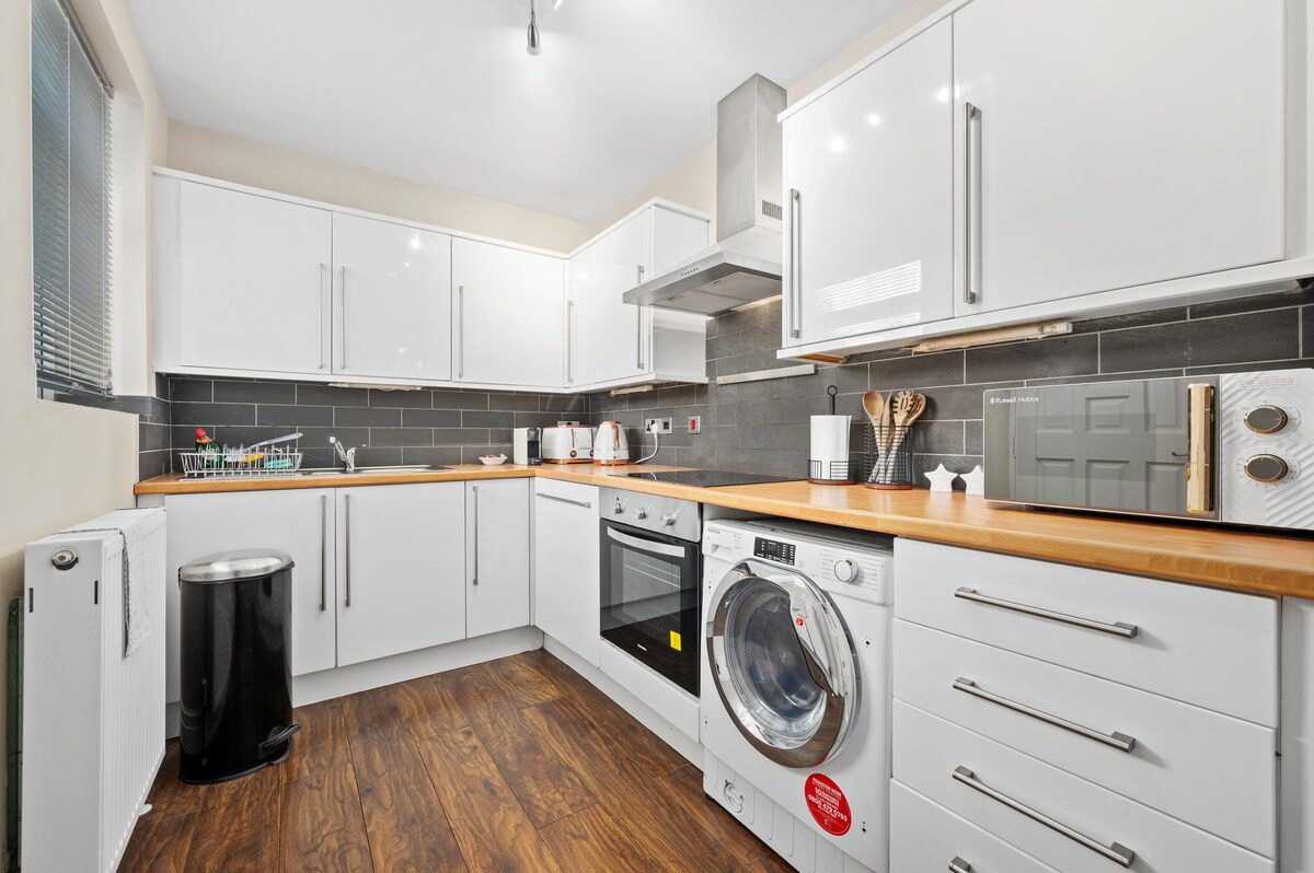 The kitchen is designed with modern cabinetry in white, featuring a mix of appliances including an oven and washing machine. A wooden countertop complements the space, while ample storage is provided through upper and lower cabinets. Large windows allow natural light to enhance the area.