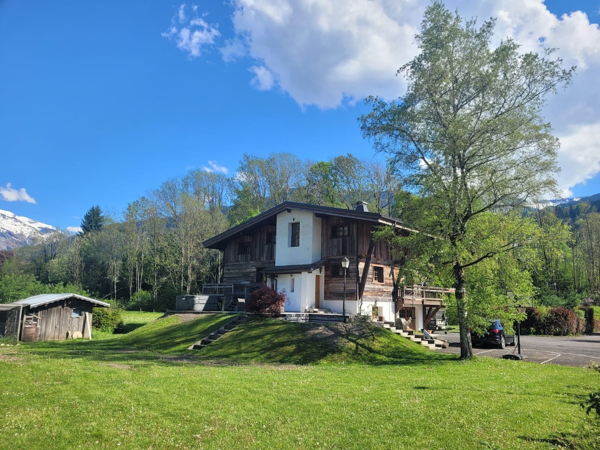A spacious alpine chalet is set against a backdrop of blue sky and greenery. The building features a combination of wood and stone materials, with a large balcony and outdoor steps leading to an inviting entrance. The surrounding landscape consists of well-maintained grass and trees.