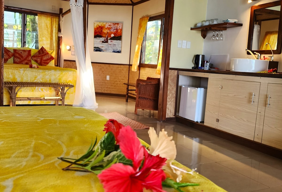 A spacious room is highlighted by warm yellow decor, featuring two beds adorned with colorful pillows. Large windows offer natural light and views, while a small kitchenette area is visible. A decorative arrangement of tropical flowers rests on the bed, contributing to the inviting ambiance.