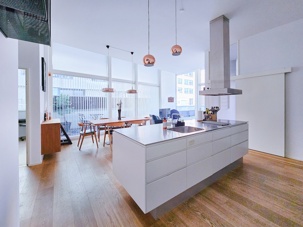 An open kitchen-dining living area features a minimalist design with a large central island and a dining table surrounded by wooden chairs. Large windows allow ample natural light, complemented by modern pendant lighting. A comfortable seating area is arranged for relaxation.
