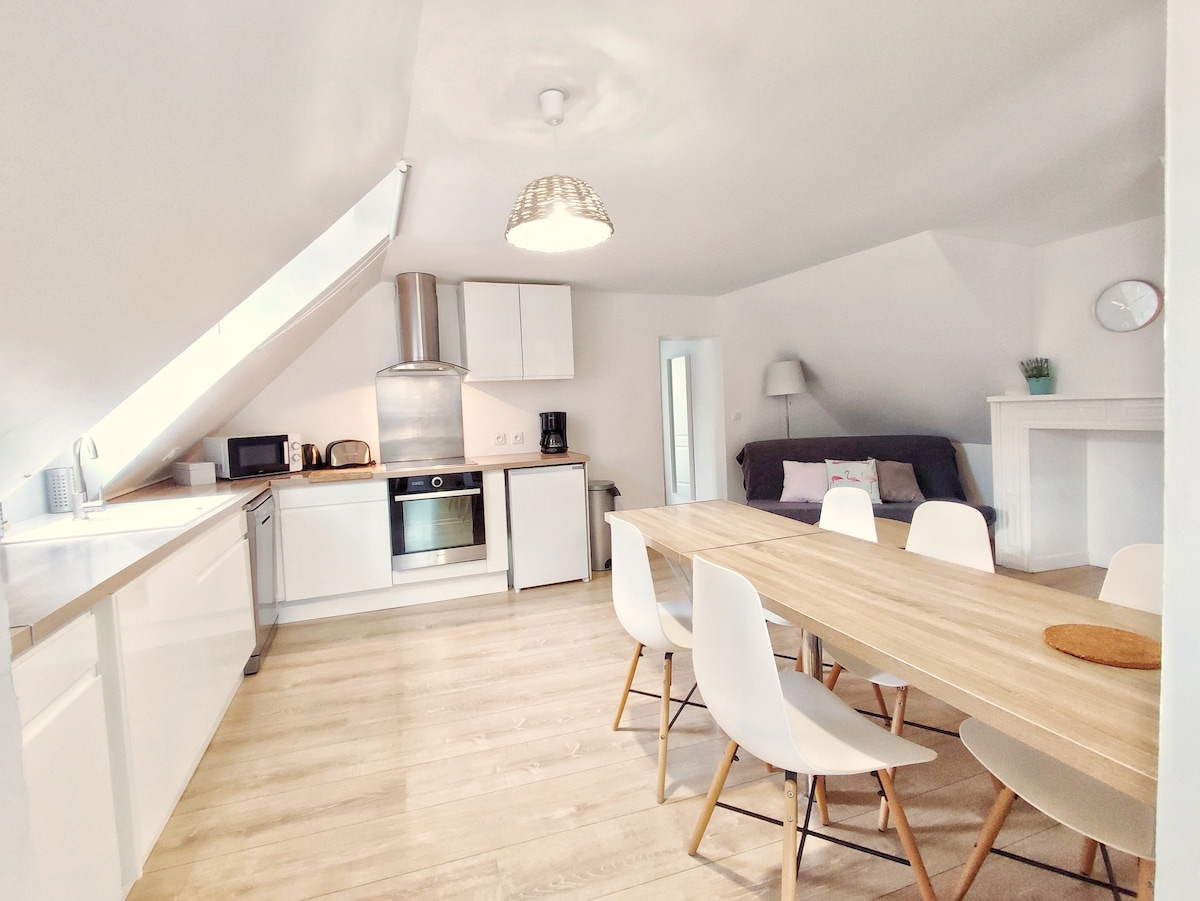 A bright kitchen and dining area features a wooden table surrounded by white chairs. Modern appliances include a stainless steel oven and hood, complemented by a minimalist layout. Natural light enters through a slanted ceiling, creating an airy atmosphere. A cozy seating area is set in a corner.