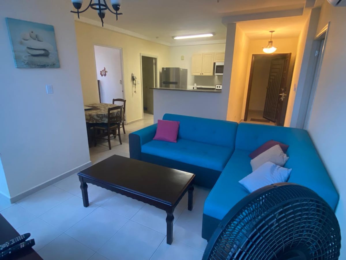 A bright living area features a turquoise sofa arranged around a dark wood coffee table. A large ceiling fan provides airflow. The space includes a dining area in the background, and the kitchen is visible with modern appliances. Light walls enhance the overall openness.
