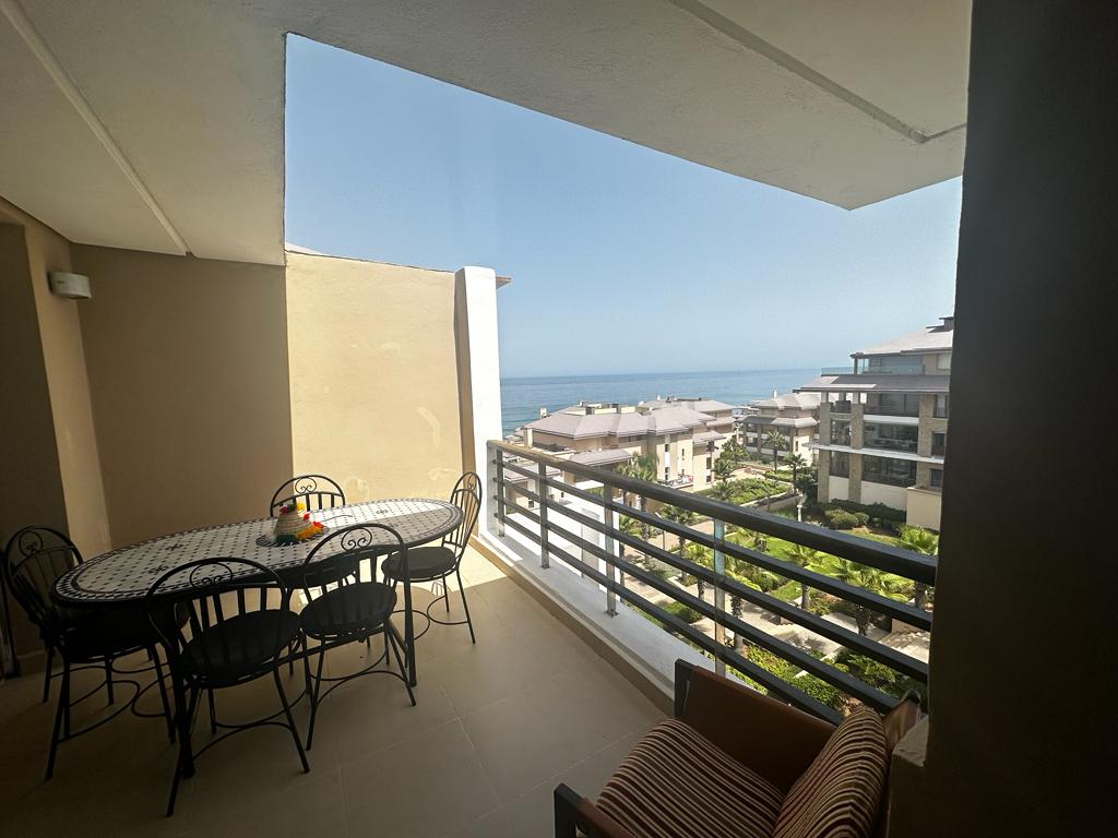 A spacious terrace is visible, featuring a round dining table surrounded by four chairs. Lush greenery and ocean views can be seen in the background, framed by a modern railing. Natural light fills the area, highlighting the inviting outdoor space.