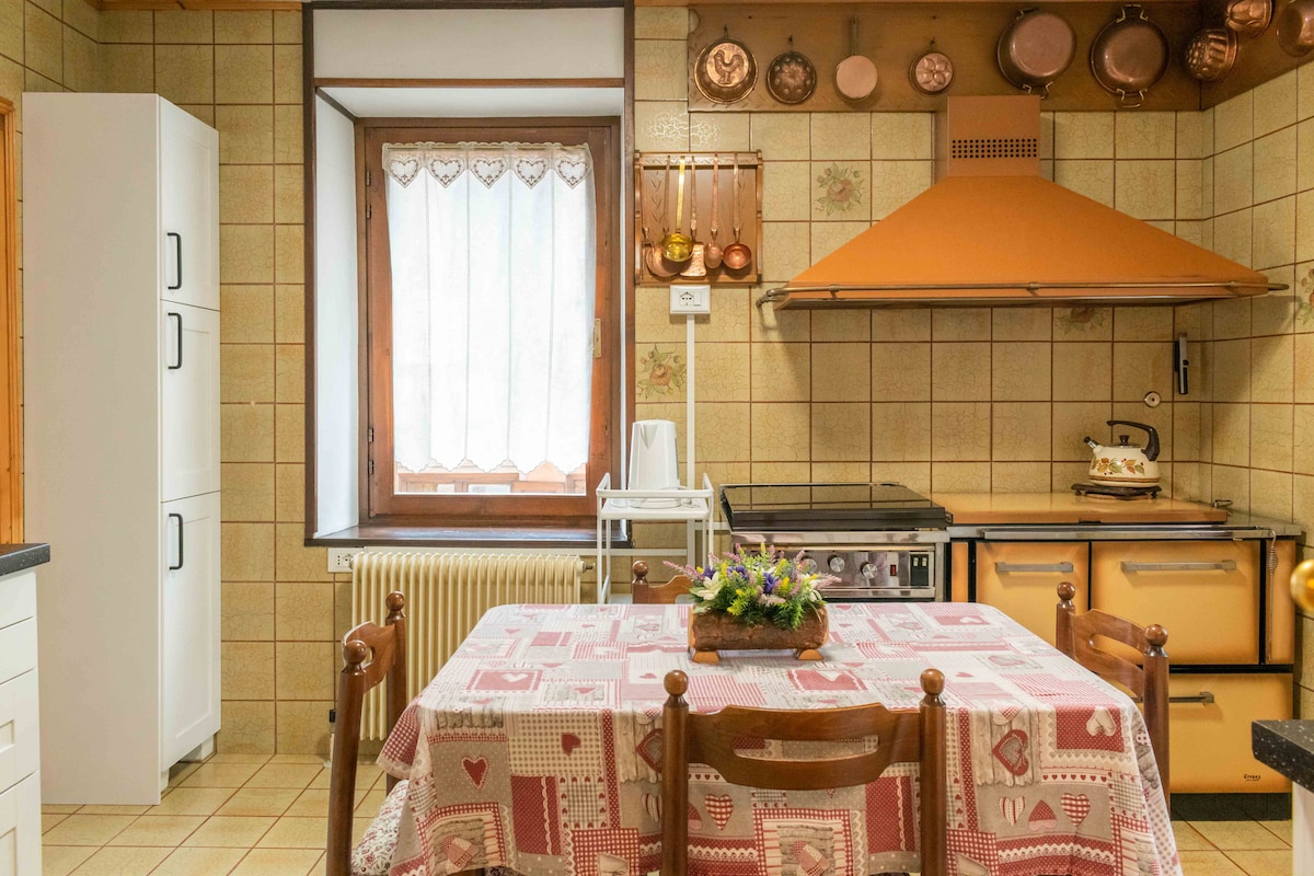 A functional kitchen space features a patterned tablecloth covering a wooden dining table with four chairs. Copper cookware hangs on the wall, complemented by a sleek orange hood above the stove. Natural light enters through a window with lace curtains, illuminating the tiled walls.