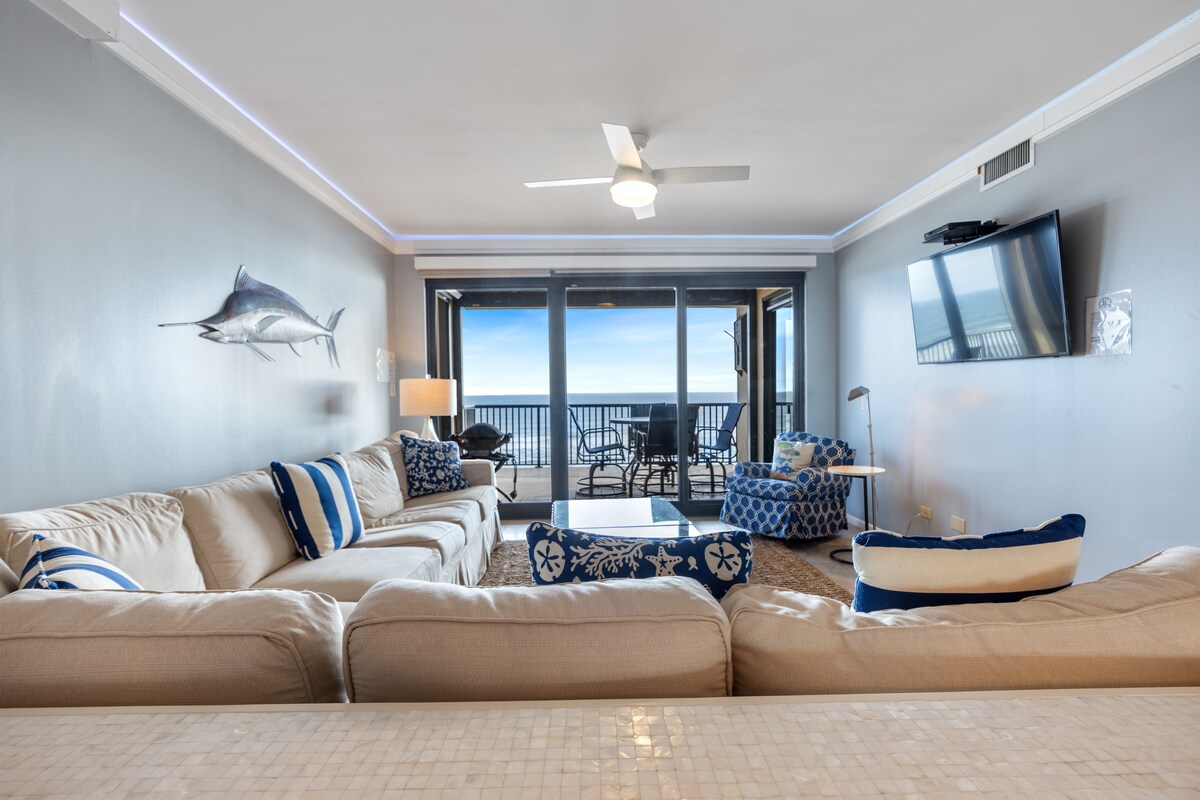 A spacious living area is furnished with a large, light-colored sectional sofa adorned with blue and white throw pillows. A modern ceiling fan hangs above, while large glass doors provide a panoramic view of the Gulf and access to the balcony.
