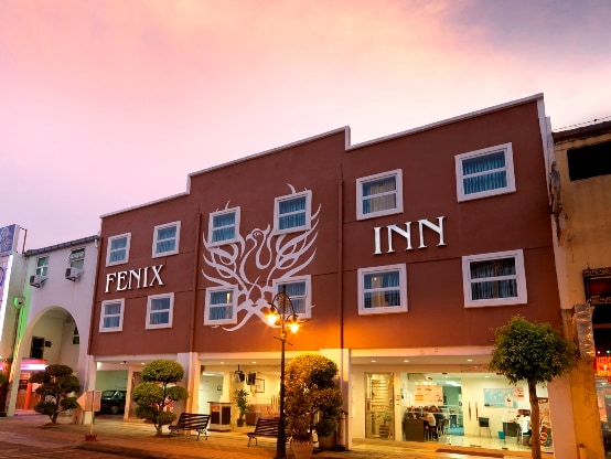 The exterior of Fenix Inn is highlighted by modern architecture and a large, welcoming sign. Soft pastel hues fill the sky, creating a serene backdrop. Symmetrical windows are flanked by neatly trimmed shrubs, enhancing the inviting entrance and providing a glimpse of the reception area.