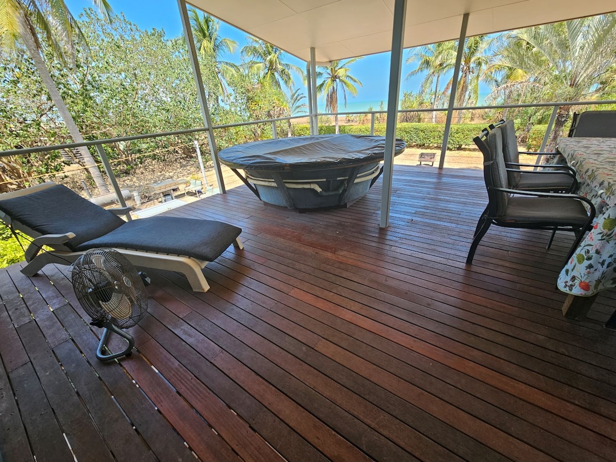 A spacious deck features a covered spa, surrounded by wooden flooring. Lounge chairs are positioned nearby, offering a relaxing spot. A dining table with chairs is visible, with greenery and ocean views seen beyond the railing. A fan is placed on the deck for comfort.
