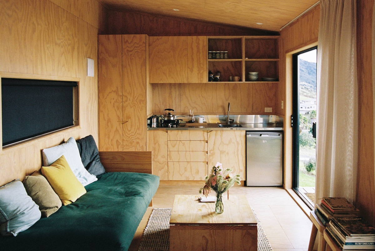 An open-plan living area features warm wooden paneling with a comfortable seating arrangement. A small table is set in the center, surrounded by books. The kitchen area includes modern appliances and storage, while large glass doors provide access to outdoor views.