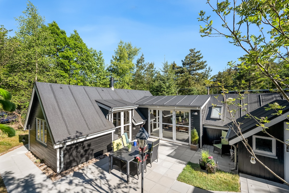 The image captures a newly renovated wooden summer house surrounded by trees. A spacious patio area is visible, featuring a dining table and chairs. Large glass doors allow natural light into the interior, which harmonizes with the outdoor scenery.