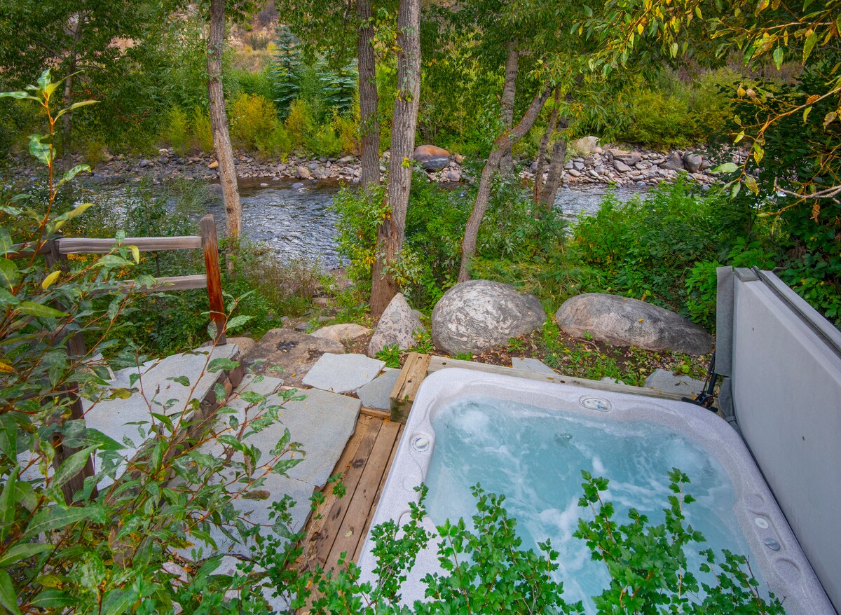 A private hot tub is situated on the edge of the river, surrounded by lush greenery and natural boulders. The patio area is composed of wooden decking and stone paving, providing a serene setting to relax while enjoying the calming sounds of the flowing Eagle River.