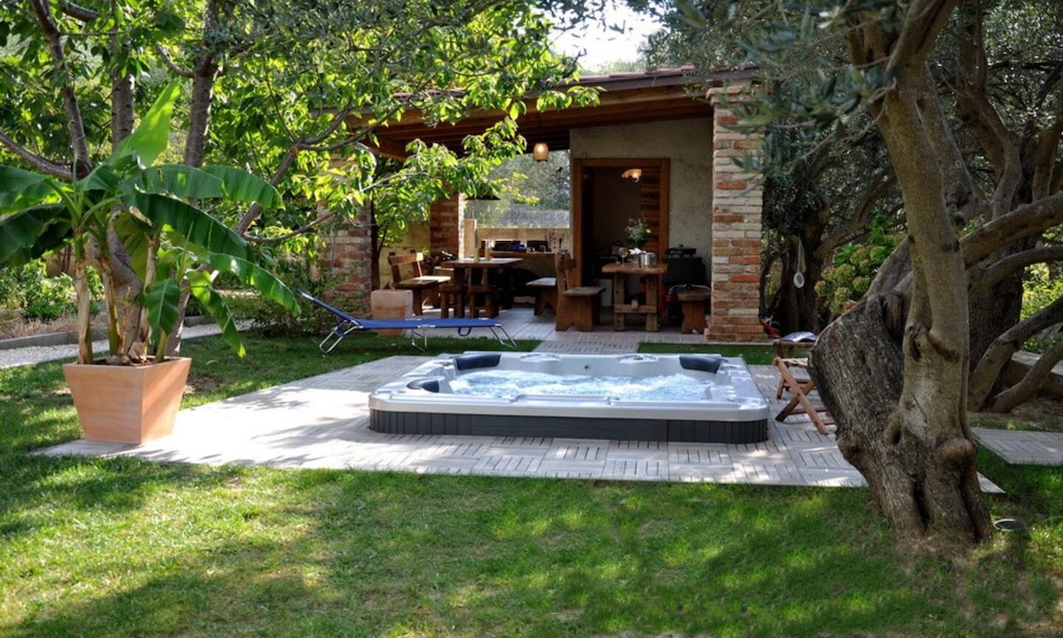 A private garden area features a hot tub surrounded by lush greenery and vibrant plants. A covered outdoor seating area with wooden furniture is visible, offering a relaxed space for dining or leisure. Sunlight filters through the trees, creating a serene atmosphere.