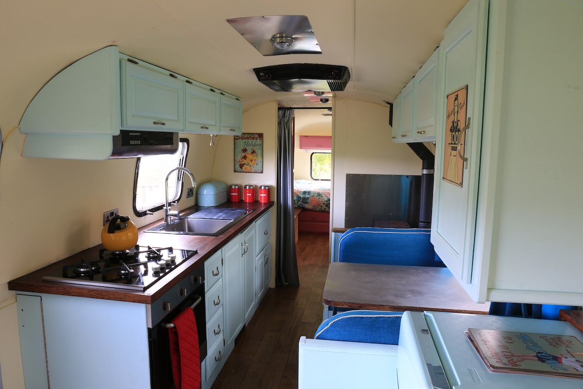 A bright kitchen area is equipped with light blue cabinetry, a sink, and a gas stove. Storage space is provided by upper and lower cabinets. A dining table with blue seating is visible, along with a small refrigerator, contributing to the functional layout of the living space.