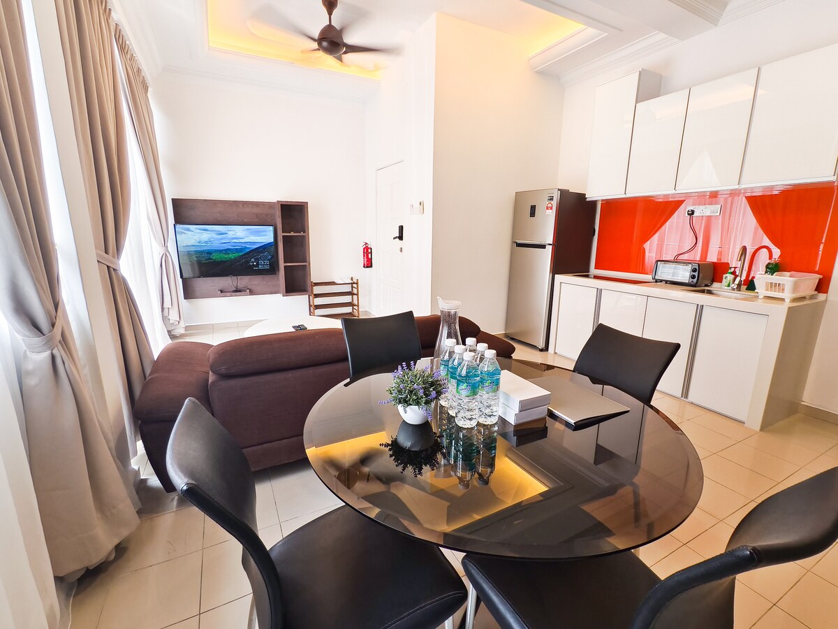 A dining area is shown with a round table set for six, accompanied by black chairs. In the background, a modern kitchen features sleek cabinetry and stainless steel appliances. A comfortable sofa is positioned nearby, with a large smart TV mounted on the wall, providing entertainment options.