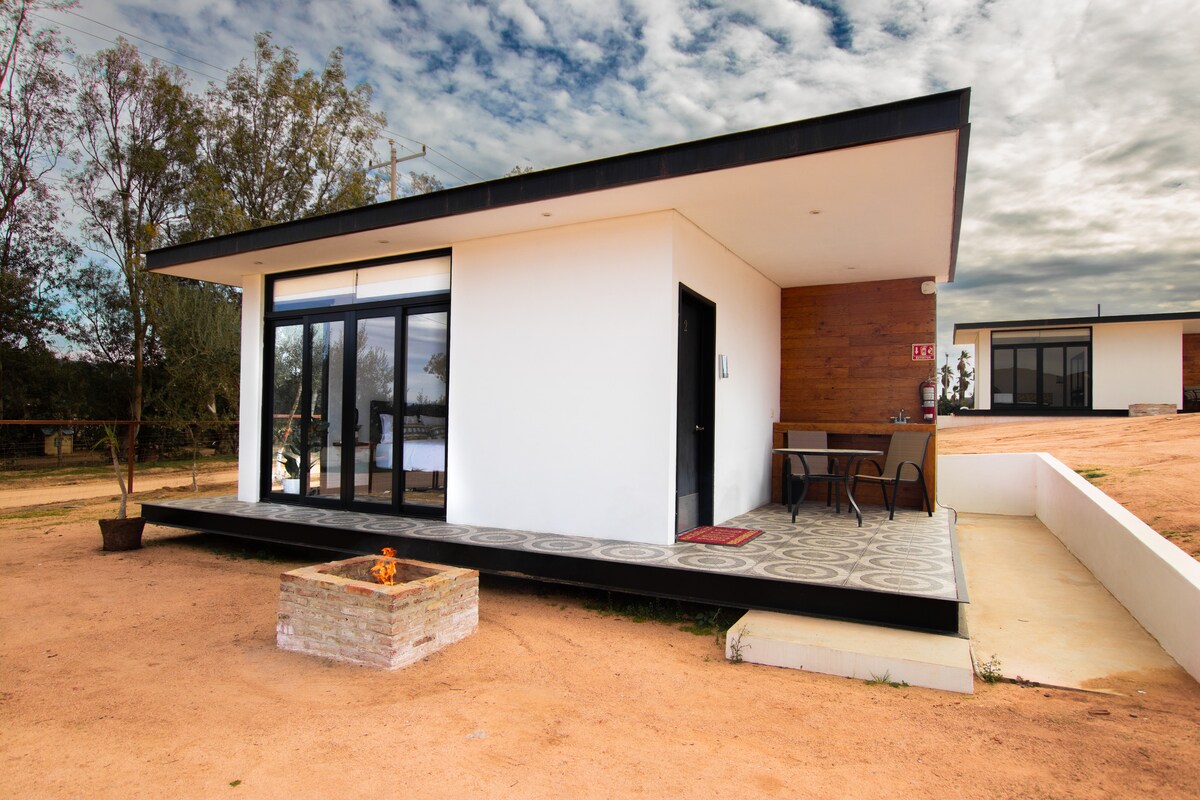 A modern building features a sleek, flat roof and expansive glass doors, providing a seamless view of the surroundings. A small outdoor seating area with a table and chairs is present on a patterned tile patio, which includes a fire pit for evening gatherings.