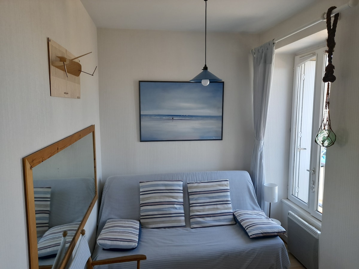 A light-filled corner of the living room features a comfortable sofa adorned with striped cushions. A wall mirror reflects the space, while a painting of a serene seascape hangs above. Natural light filters through a window dressed with a sheer curtain.