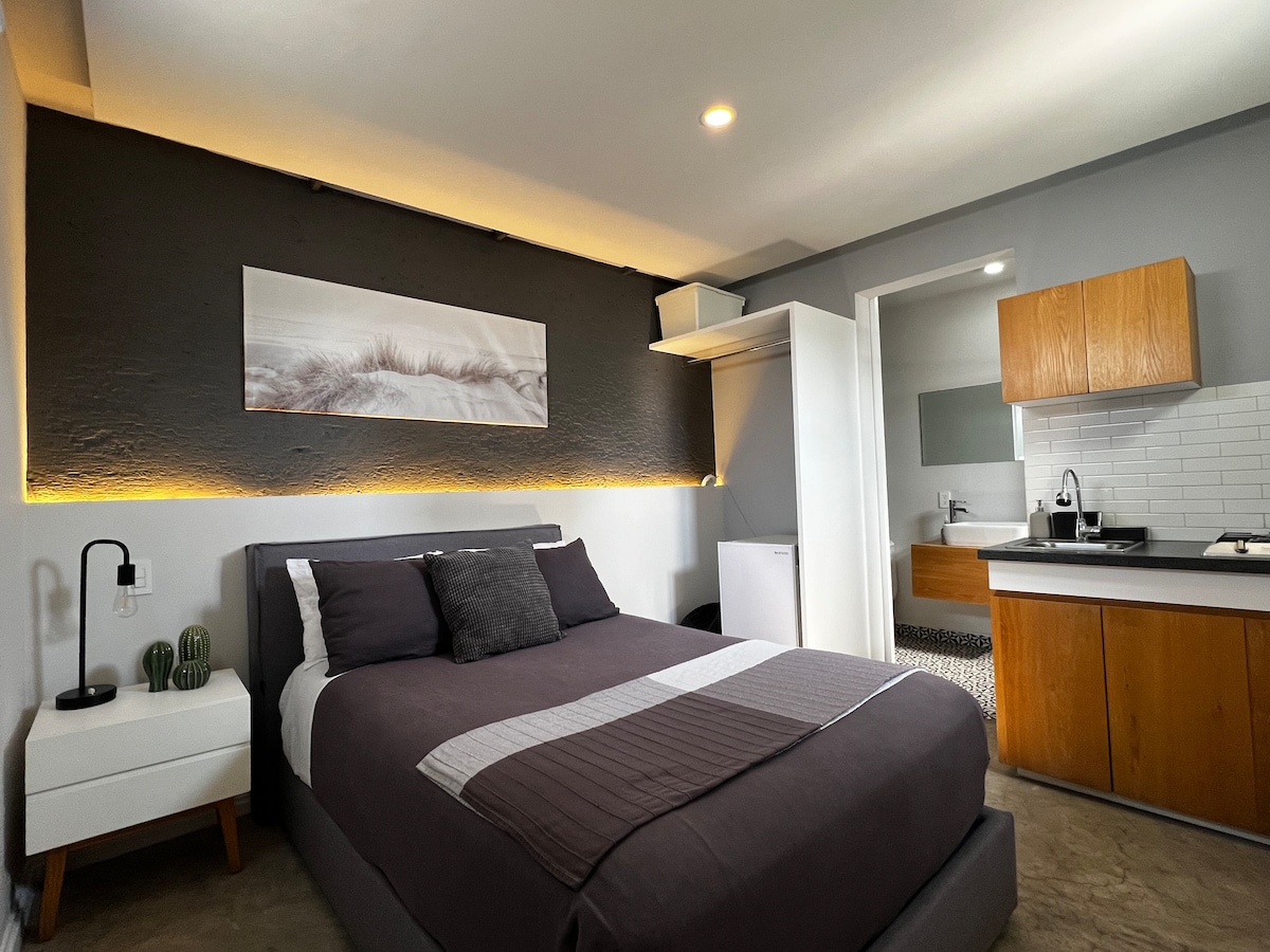 A spacious sleeping area features a comfortable bed with soft bedding. A kitchenette is visible, complemented by wooden cabinetry. Light from a wall fixture softly highlights the textured wall behind the bed, creating an inviting atmosphere. A bathroom and closet area can be seen in the background.