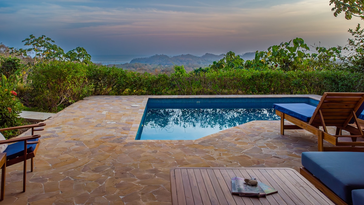 An inviting outdoor area features a clear swimming pool surrounded by natural stone flooring. Comfortable lounge chairs offer a place to relax, with lush greenery framing the view of distant hills and the ocean beyond, all under a softly lit sky.