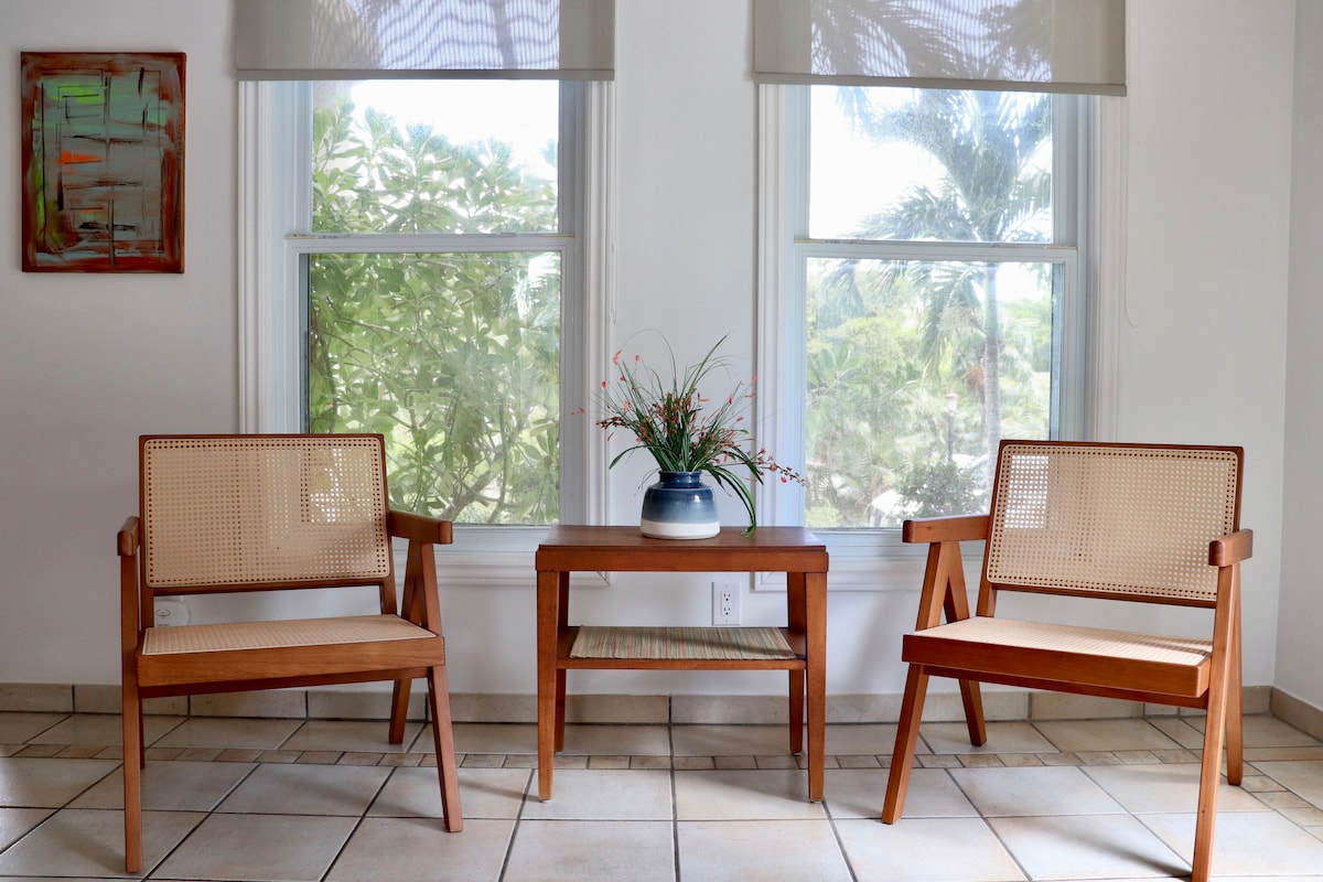 A seating area is depicted with two wooden chairs featuring cane backs and light cushions. A small wooden table is positioned between them, topped with a vase and greenery. Natural light filters through two large windows, showcasing lush foliage outside.