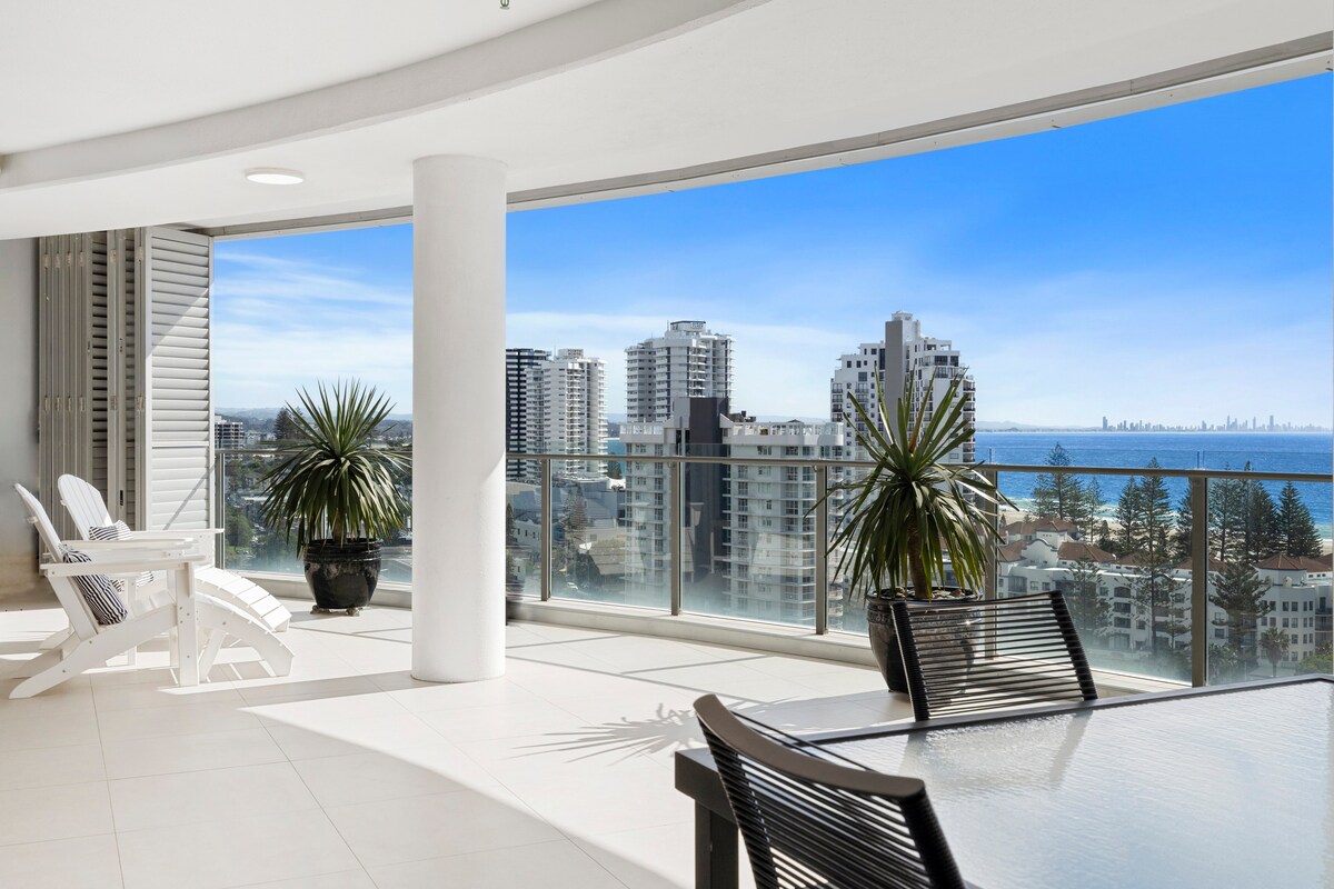 An expansive balcony is featured, providing sweeping views of the coastline and city skyline. The area includes outdoor seating with a glass-top table and comfortable lounge chairs. Glass railings enhance the openness, while potted greenery adds a touch of nature to the space.