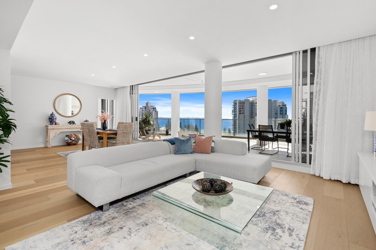 A spacious living area features a modern sectional sofa and a glass coffee table atop a soft rug. Floor-to-ceiling windows provide a view of the ocean, while natural light fills the room. A dining area with woven chairs is visible in the background.