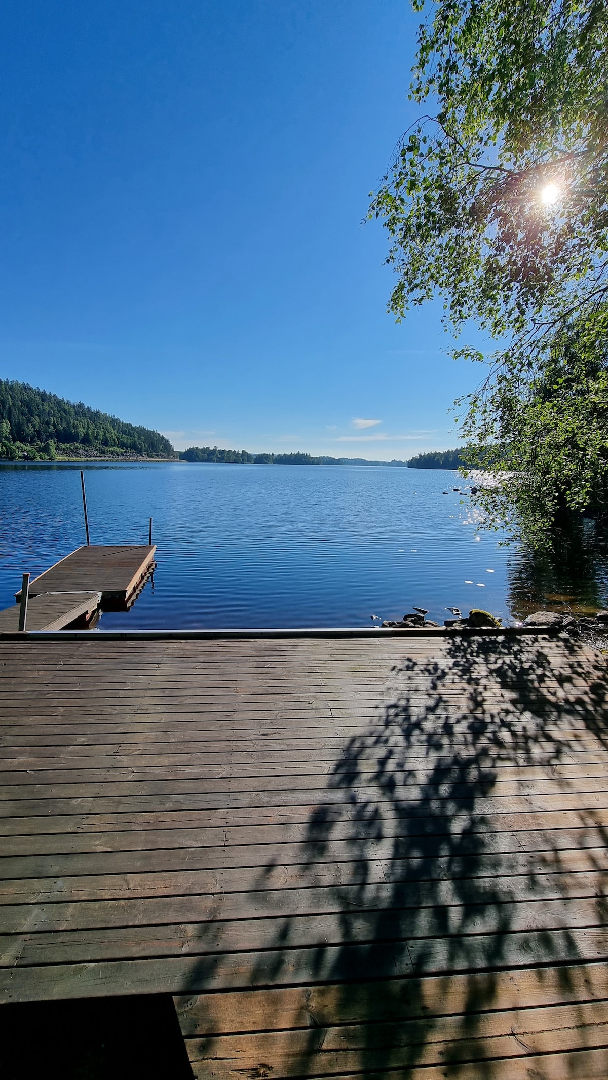 A serene lake view is presented, showcasing calm waters reflecting a clear blue sky. A wooden dock extends into the lake, bordered by gentle greenery on the shore. Dappled sunlight casts soft shadows on the wooden surface, enhancing the tranquil atmosphere.