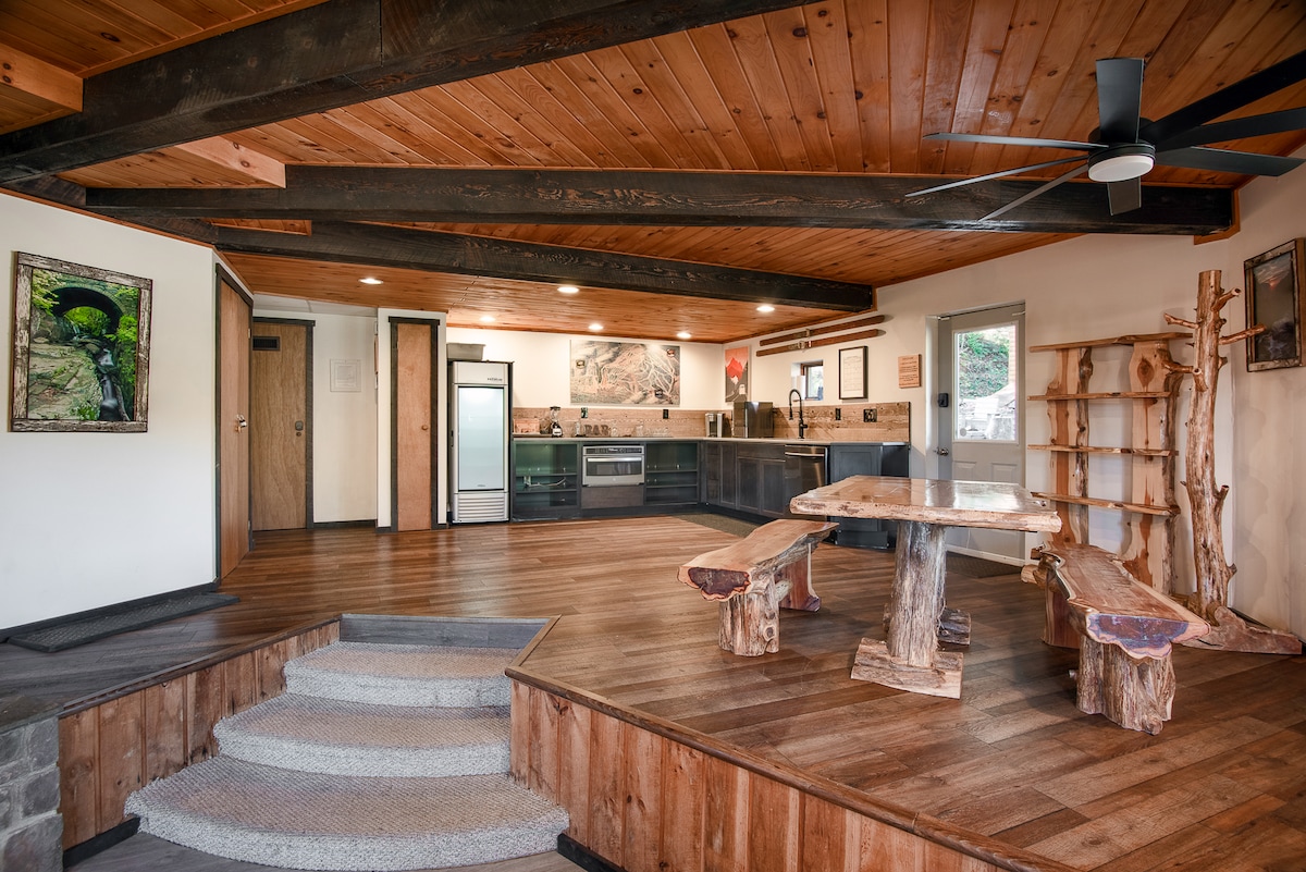 An open kitchen area with wooden beams and a rustic design is featured. The space includes a spacious dining table made of natural wood, complemented by stone and wood accents. Large windows allow soft light to fill the room, highlighting the warm tones of the wooden flooring.