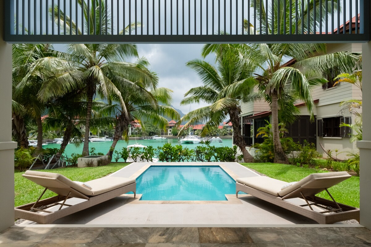 A serene outdoor area showcases a small pool surrounded by lush greenery and palm trees. Two lounge chairs are positioned beside the pool, offering a view of the water. The environment is tranquil, with light filtering through the leaves.