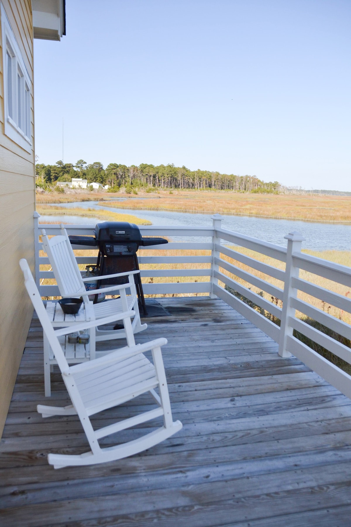 A wooden deck features two white rocking chairs positioned for serene views. A black gas grill is seen nearby, with a landscape of marshland extending to the horizon, showcasing natural beauty and a sense of tranquility.