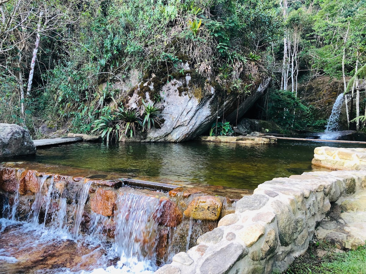 A natural swimming pool is surrounded by lush greenery, with a gentle waterfall cascading over stone. Large boulders are partially covered in moss, and the water reflects the colors of the surrounding foliage, creating a serene environment in a peaceful outdoor setting.