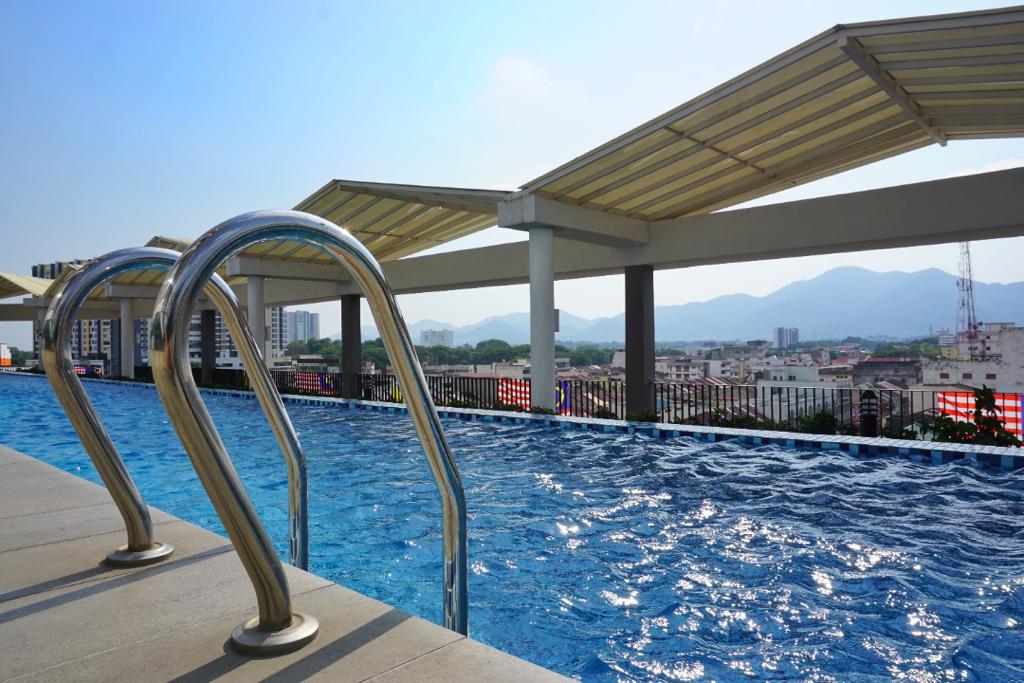 A rooftop pool is visible, featuring a smooth surface of sparkling blue water. Two stainless steel handrails lead into the pool area, with a backdrop of distant mountains and city buildings under a clear sky.