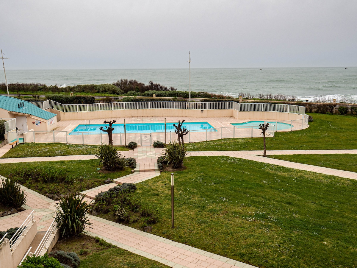 An outdoor heated swimming pool is situated adjacent to the ocean, surrounded by well-maintained gardens. A pathway leads to the pool area, with lounge chairs positioned for relaxation. Lush greenery and decorative plants enhance the tranquil environment, creating a scenic view of the coastline.
