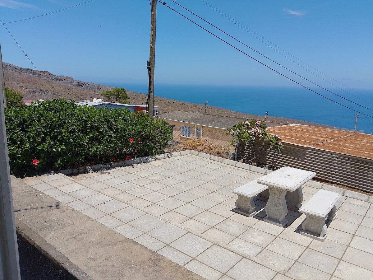 An outdoor patio area features a paved surface complemented by low greenery and flowering plants. A stone table with matching benches is centrally positioned, offering a view of the expansive ocean and distant hills under a clear blue sky.