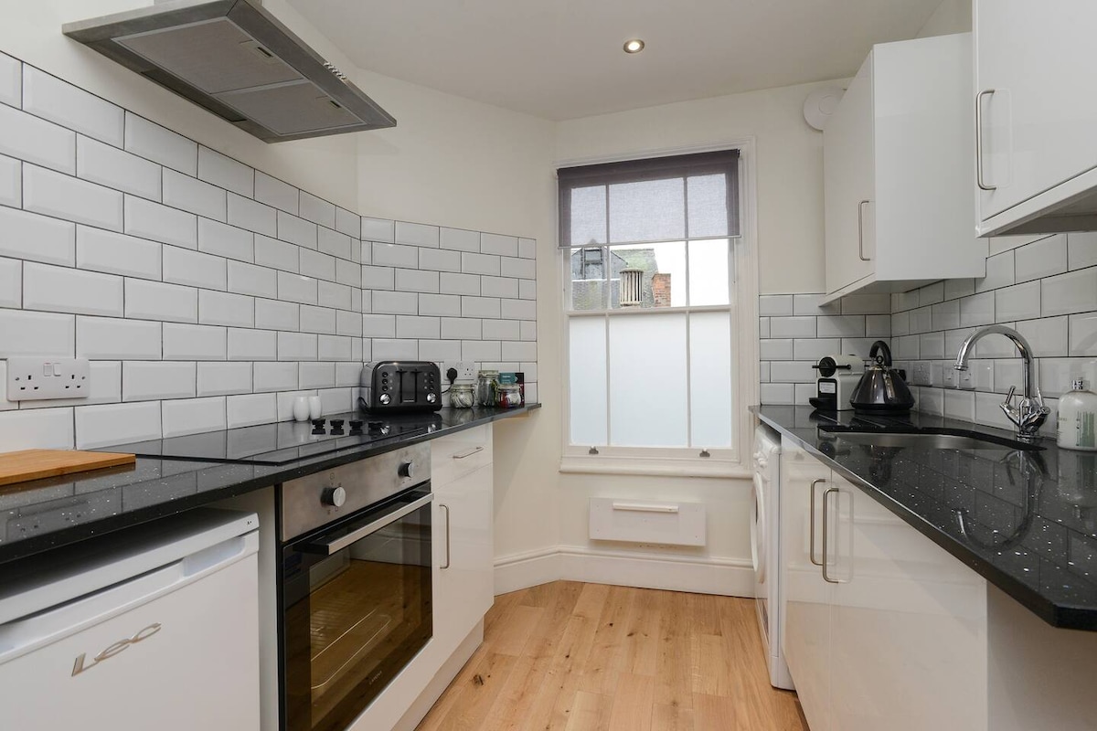 The kitchen features modern white cabinetry and black quartz countertops. A combination oven, stovetop, and kettle are visible. Subway tile backsplash complements the design. Natural light enters through a large window, partially covered by light grey blinds.