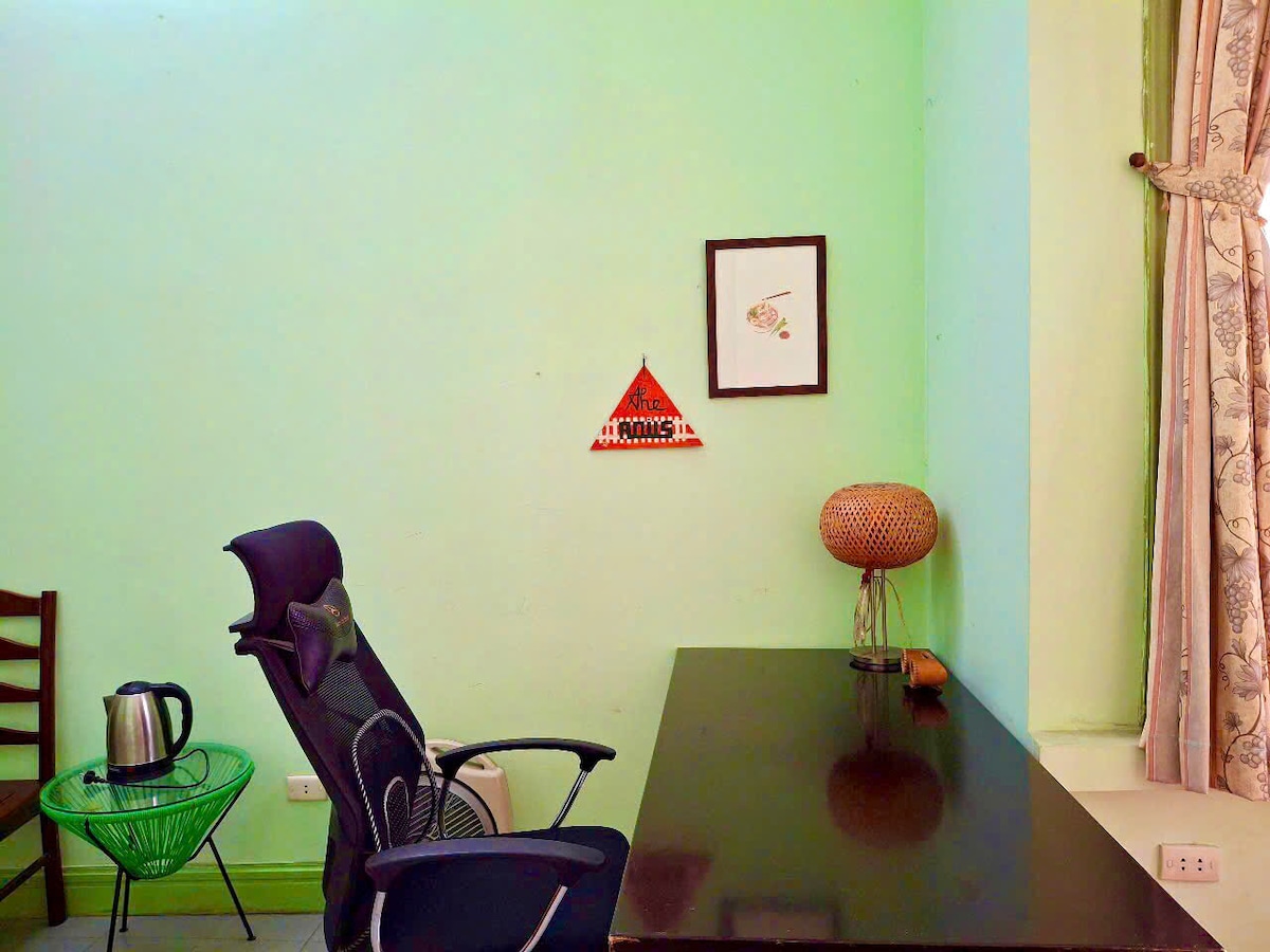 A workspace is presented with a dark wooden desk and a comfortable chair. A kettle is visible on the desk, alongside a decorative lamp. The walls are painted in soft green hues, complemented by curtains and framed artwork, providing a calm environment.