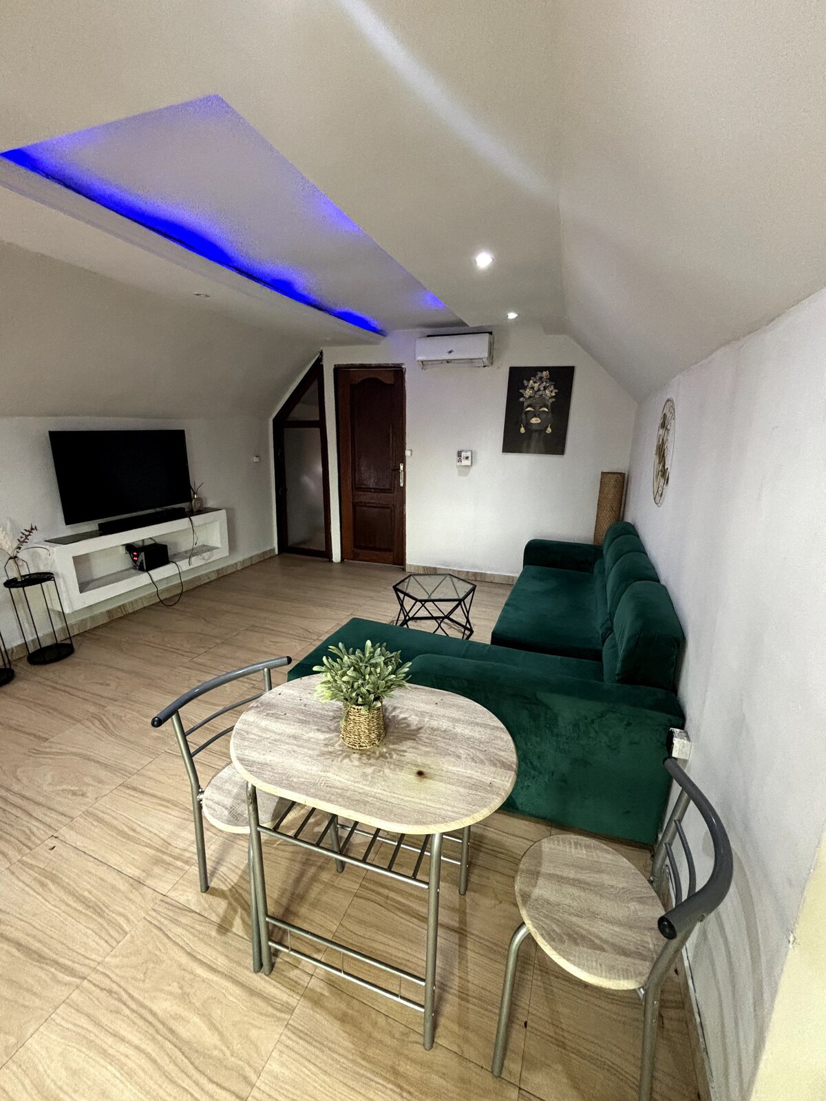 The comfortable living area features a green couch, a small table with two chairs, and a wall-mounted television. Soft lighting is provided by recessed ceiling lights, while a decorative plant adds a touch of greenery to the space.