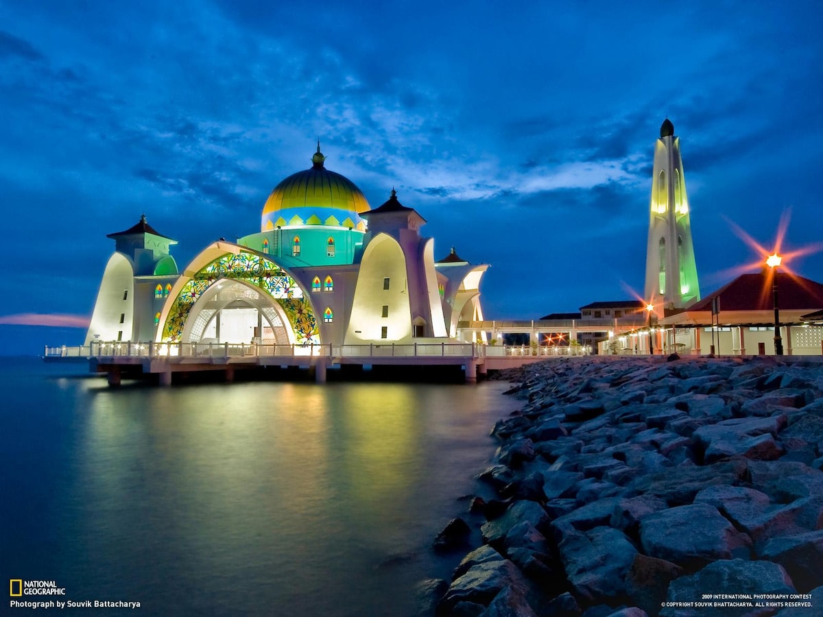 A modern mosque stands gracefully on the water, highlighted by intricate architectural details and colorful stained glass. The tall minaret is visible beside the structure, both illuminated against a twilight sky. Gentle ripples reflect the serene environment along the rocky shore.
