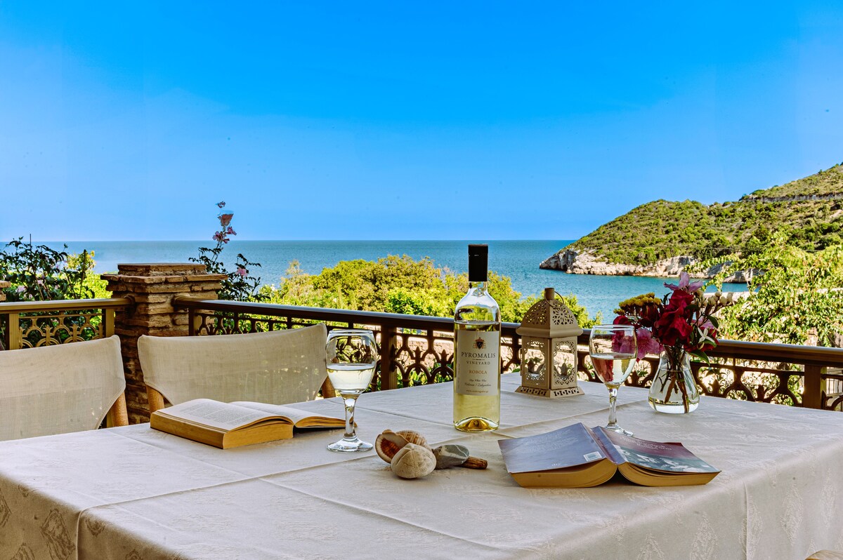 A serene outdoor dining area features a white tablecloth draped over a table set for guests. Two glasses of wine and an open bottle sit alongside books and decorative flowers, with the clear blue sea visible in the background, framed by greenery.