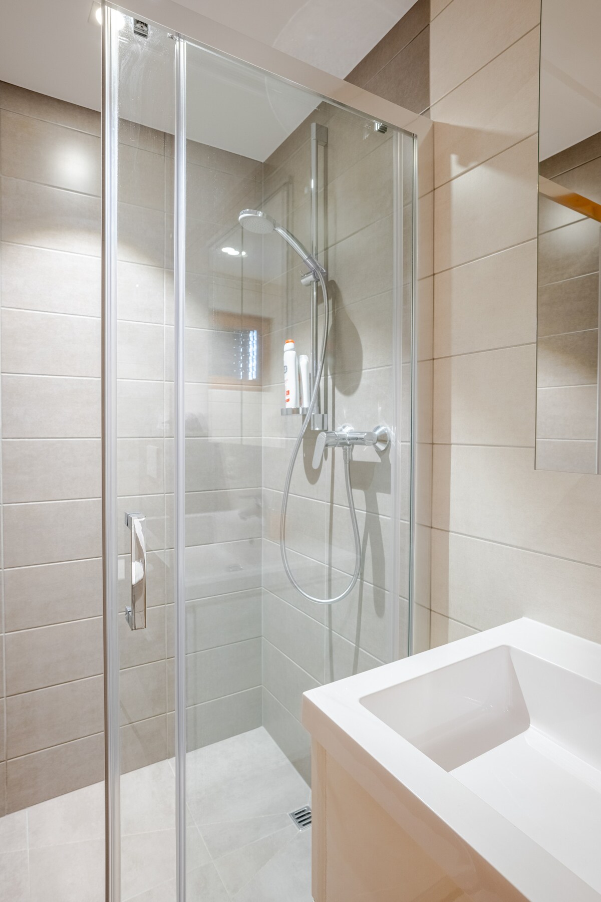 A sleek and modern bathroom is showcased, featuring a glass shower enclosure with silver fixtures. Neutral-toned tiles cover the walls, while a minimalist white sink is positioned nearby. Soft lighting enhances the clean and fresh look of the space.