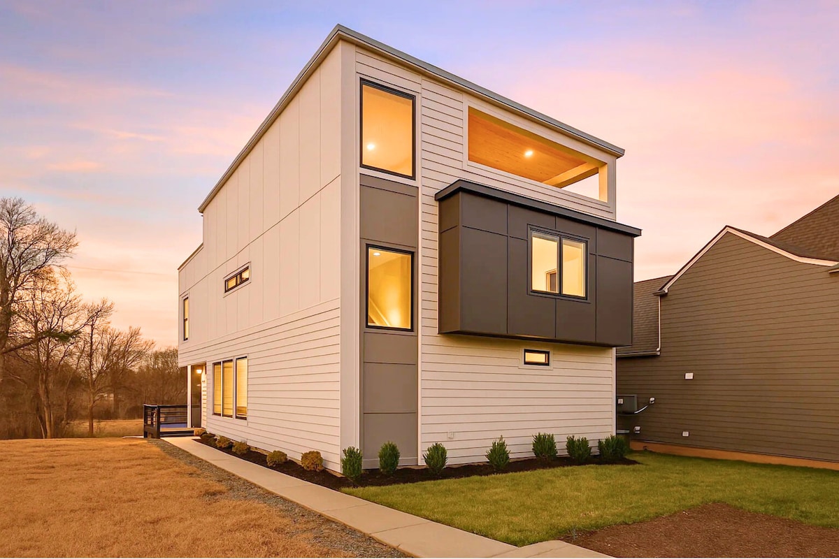 The modern home features a two-story structure with a blend of white siding and dark accents. Large windows provide ample natural light, and the front yard is landscaped with low-maintenance greenery. A gentle slope leads to a wooden deck along the side.