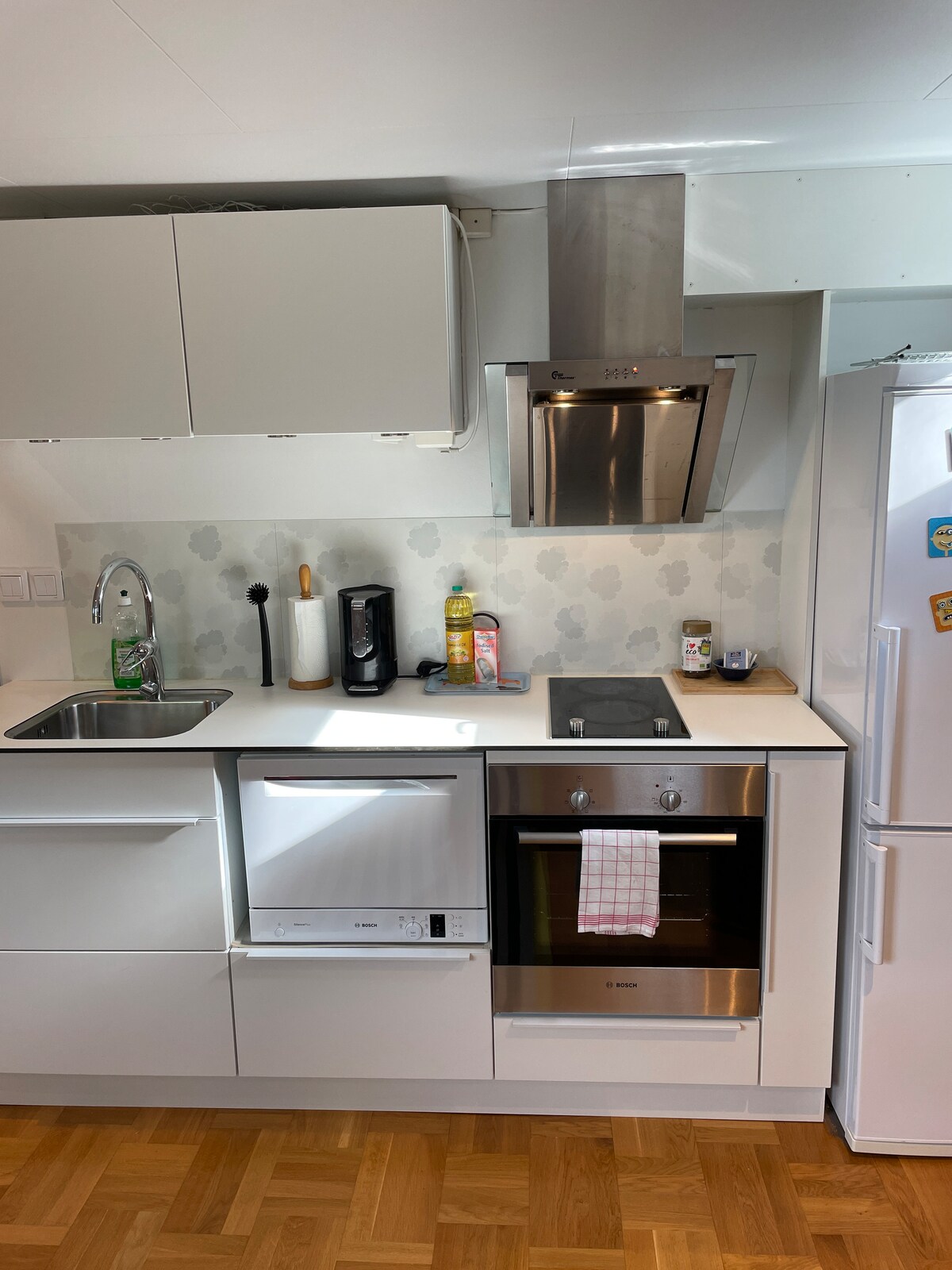The kitchen features modern white cabinetry and a sleek countertop. Equipped with a sink, stovetop, and oven, it offers essential appliances, including a dishwasher and a coffee maker. Natural light illuminates the space, creating an efficient area for meal preparation.