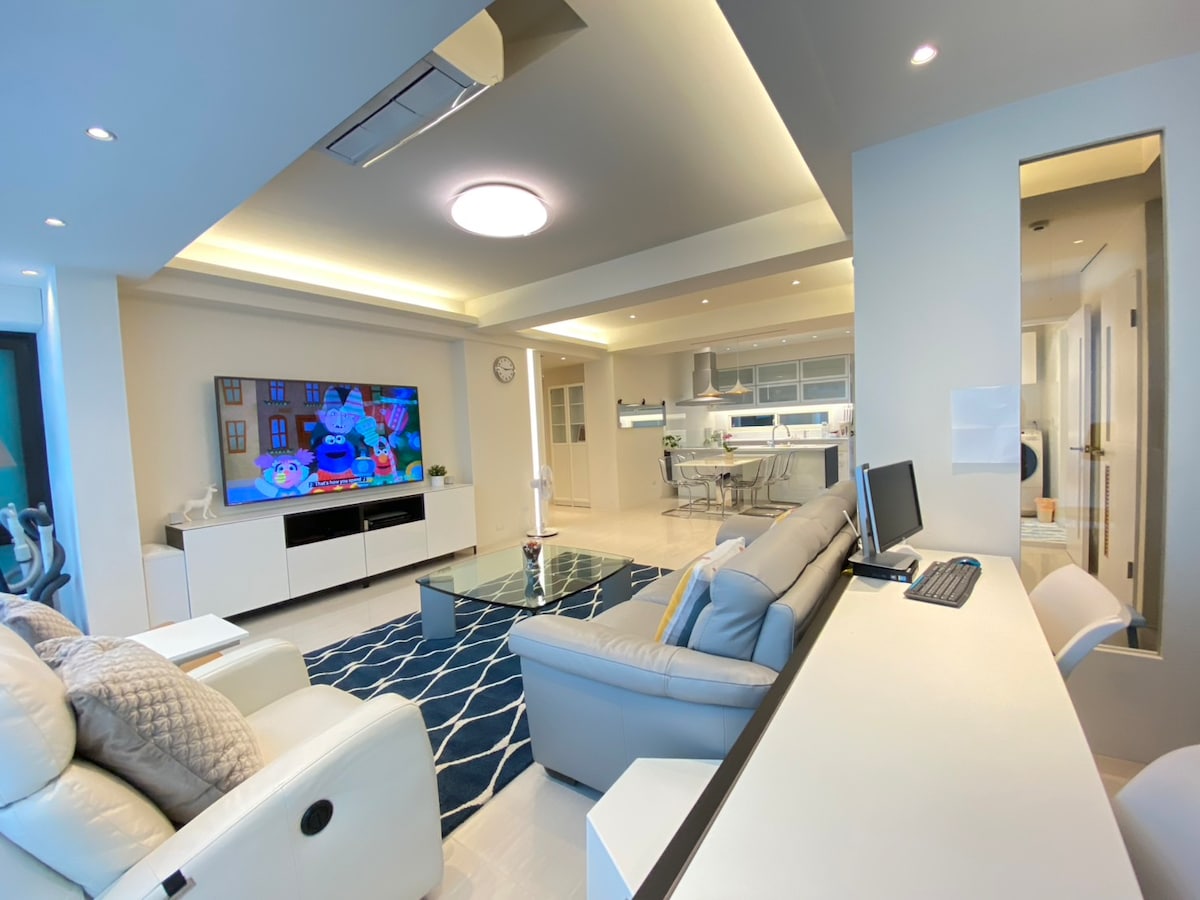 The living space is characterized by a light and airy design, featuring a large flat-screen television mounted on the wall. A cozy gray sectional sofa is positioned around a glass coffee table, while a modern dining area and kitchen are visible in the background.