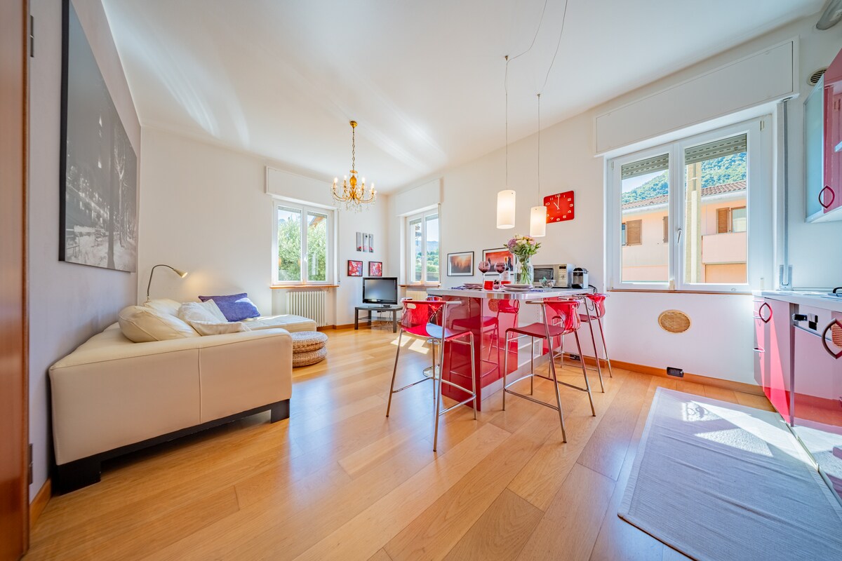 A bright open space living area is equipped with a comfortable sofa and a television, surrounded by large windows that invite natural light. A dining table with red chairs and contemporary lighting enhances the ambiance, while a well-equipped kitchen area is visible in the background.