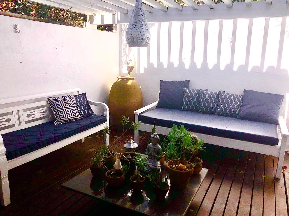 A serene outdoor seating area features two spacious sofas adorned with dark blue cushions. A glass table sits between them, surrounded by potted plants. A decorative sculpture is placed on the table, complemented by a large ceramic pot in the background and dappled sunlight filtering through the slats above.