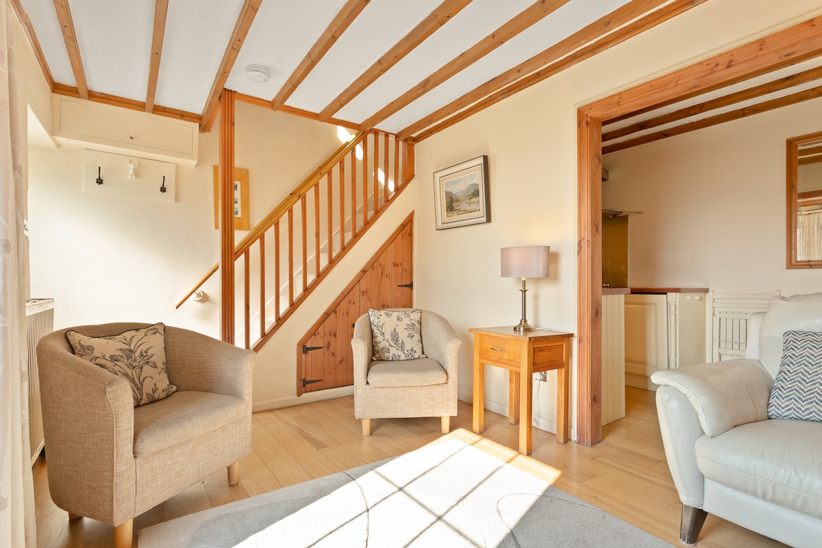 The sitting area features two light upholstered armchairs and a small wooden table, complemented by natural wood accents and a cozy carpet. A staircase leads to the upper level, while a doorway opens to a modern kitchen space in the background, allowing for seamless access between areas.