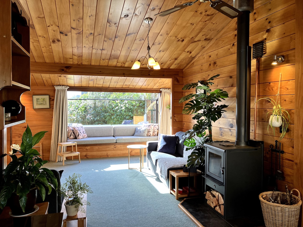 A warm living area showcases wooden walls and ceiling, complemented by a cozy seating arrangement. A wood stove is situated beside a tranquil view of greenery through large windows, while potted plants add a touch of nature to the space.