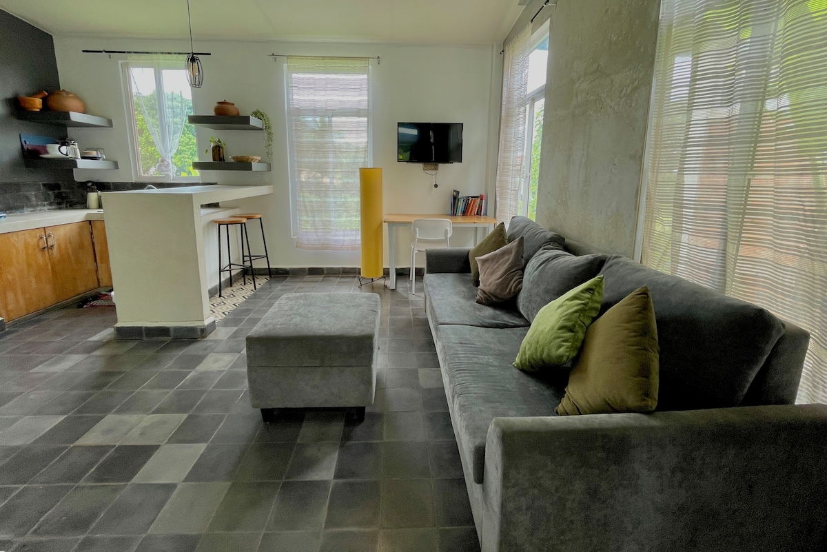 The living area features a spacious gray sofa with decorative cushions, complemented by a gray ottoman. Natural light filters through sheer window treatments, illuminating the modern kitchen with wooden cabinetry and barstools. A small dining area with a minimalist table and chair adds functionality to the space.