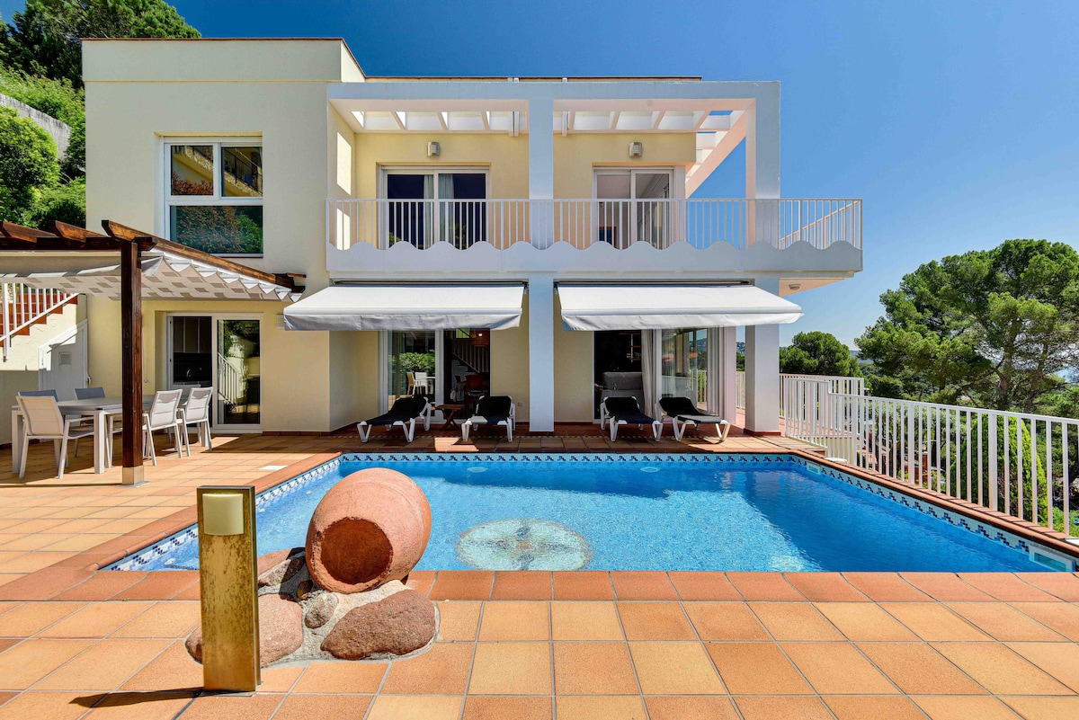 The villa features a spacious terrace with a private semi-salted pool, surrounded by sun loungers. Large glass doors lead indoors, while retractable awnings provide shade over the outdoor dining area. The vibrant blue pool contrasts against the warm, tiled flooring and lush greenery.